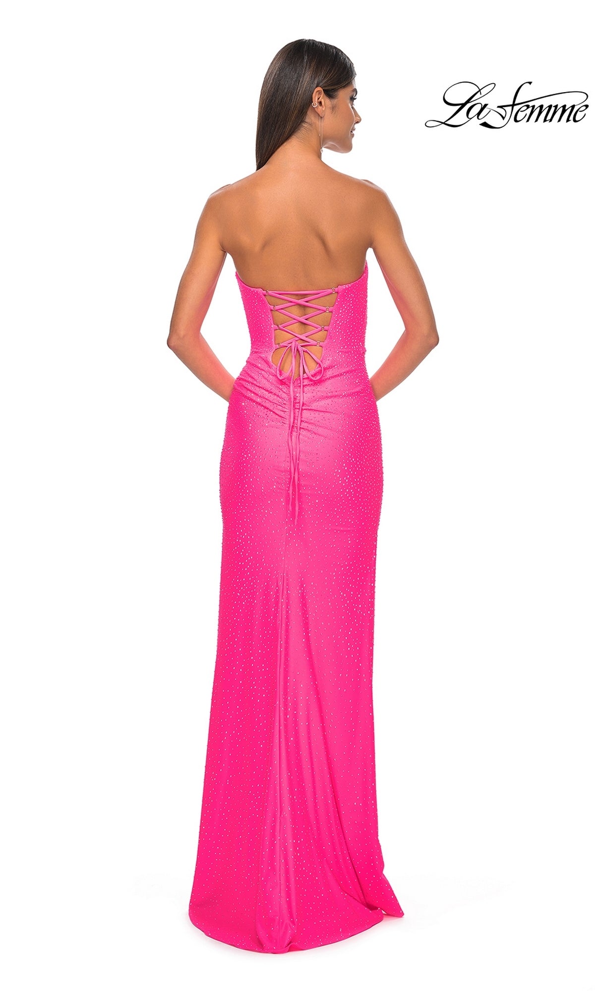 A woman stands facing away in the La Femme Twist Strapless Long Prom Dress 32175—a bright pink, floor-length gown with a lace-up back, fitted silhouette, and subtle sparkling embellishments.